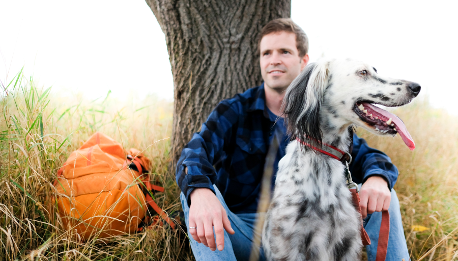 Ein Mann sitzt vor einem Baum mit seinem orangefarbenen Rucksack auf der Wiese und sein Hund sitzt vor ihm auf einem Blatt